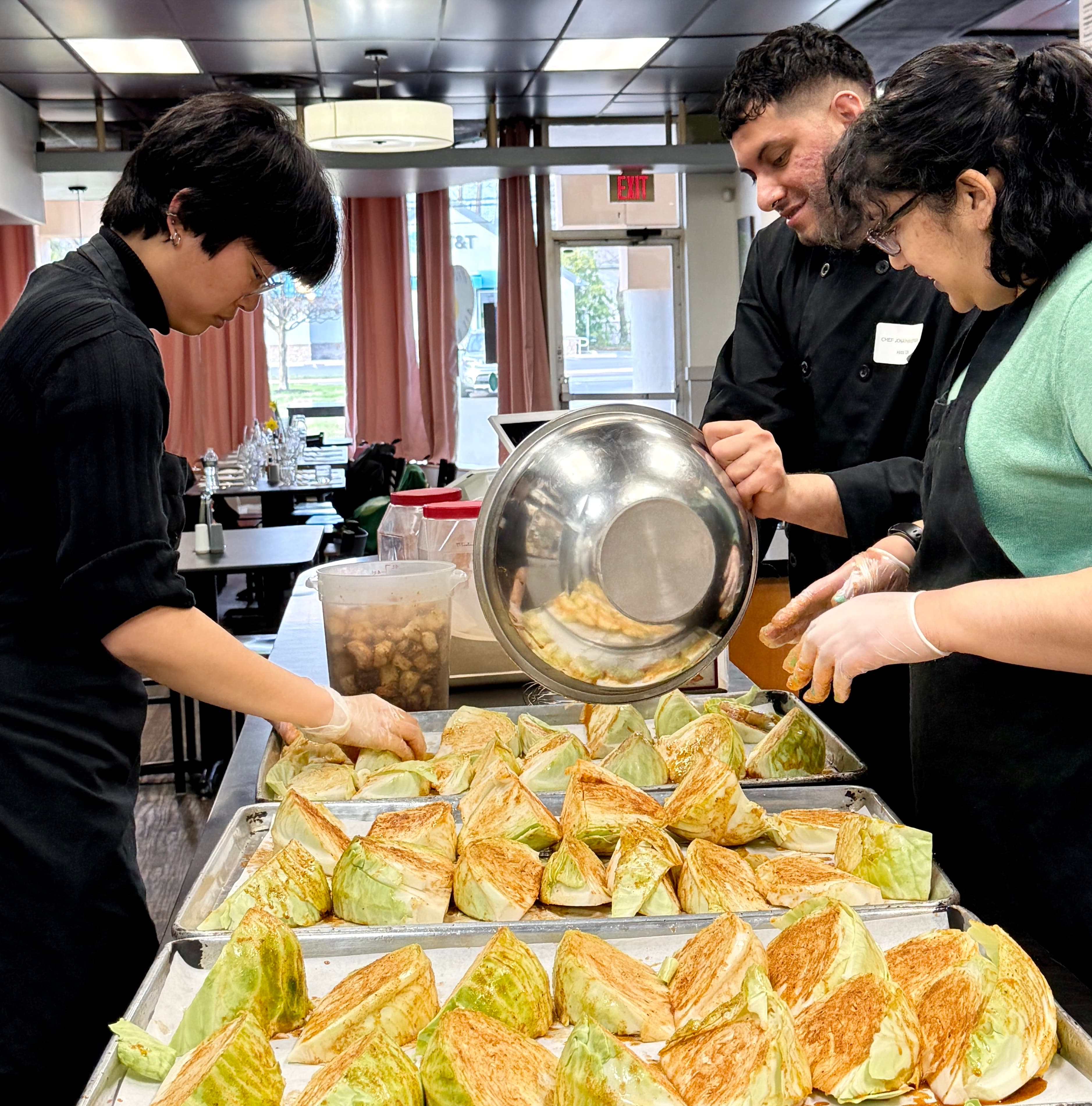 Three people in a kitchen preparing large trays of seasoned cabbage wedges for roasting.