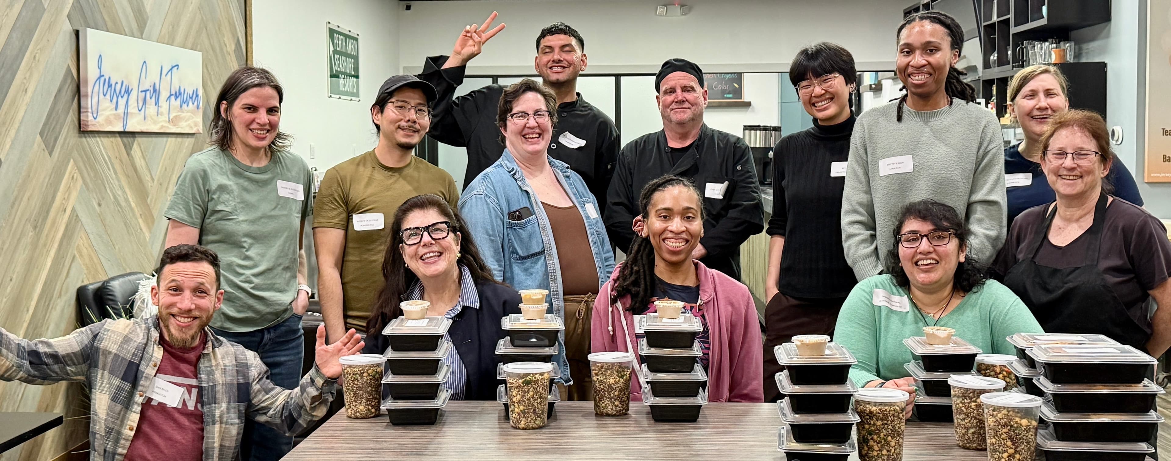 Diverse group of smiling people posing behind stacks of prepared meal containers and jars.