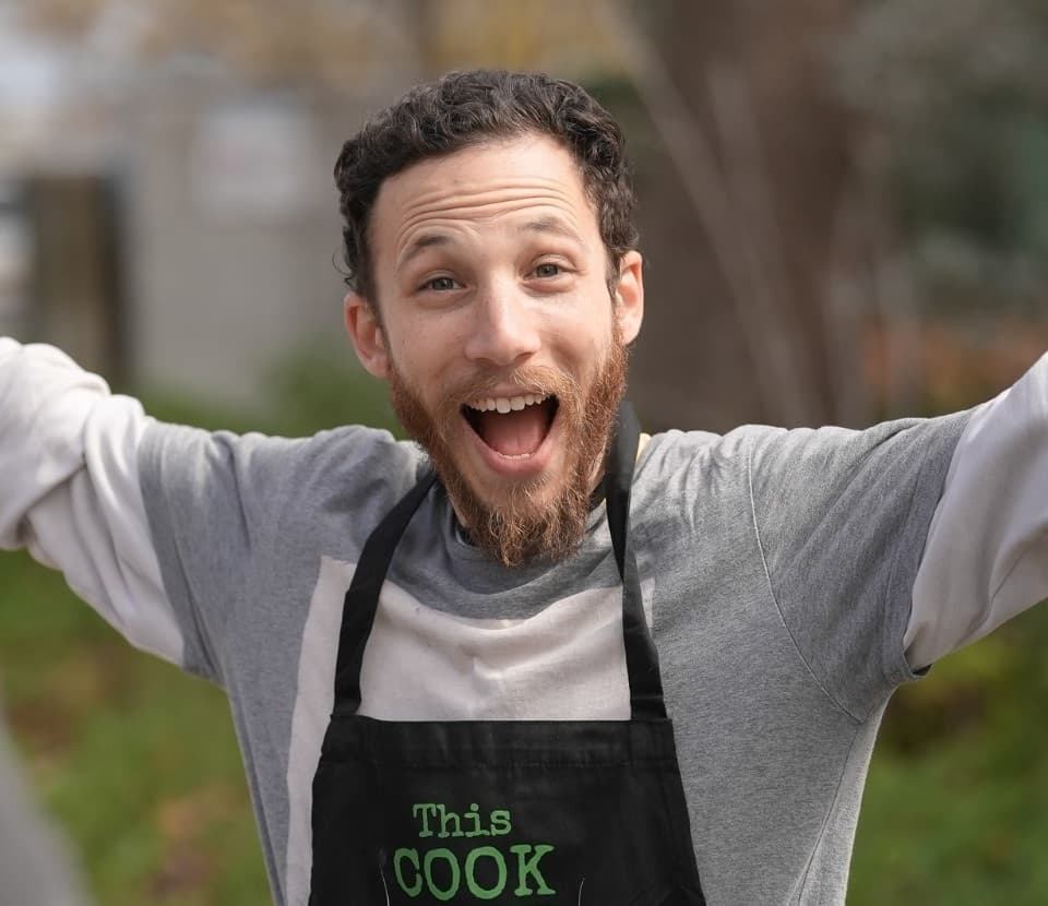 Bearded man wearing a "This COOK" apron smiles excitedly with his arms raised outdoors.