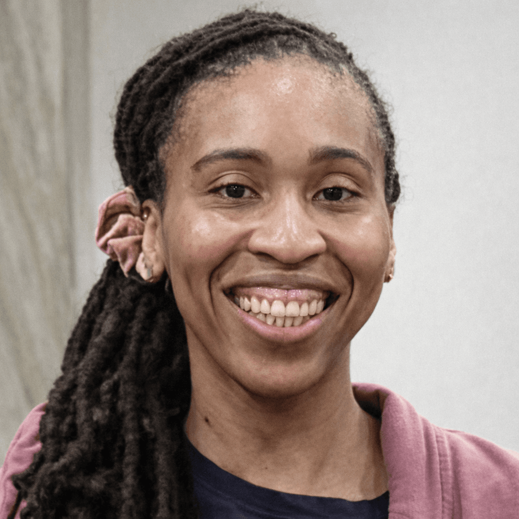 Smiling Black woman with long dreadlocks tied back, wearing a pink cardigan and blue shirt.
