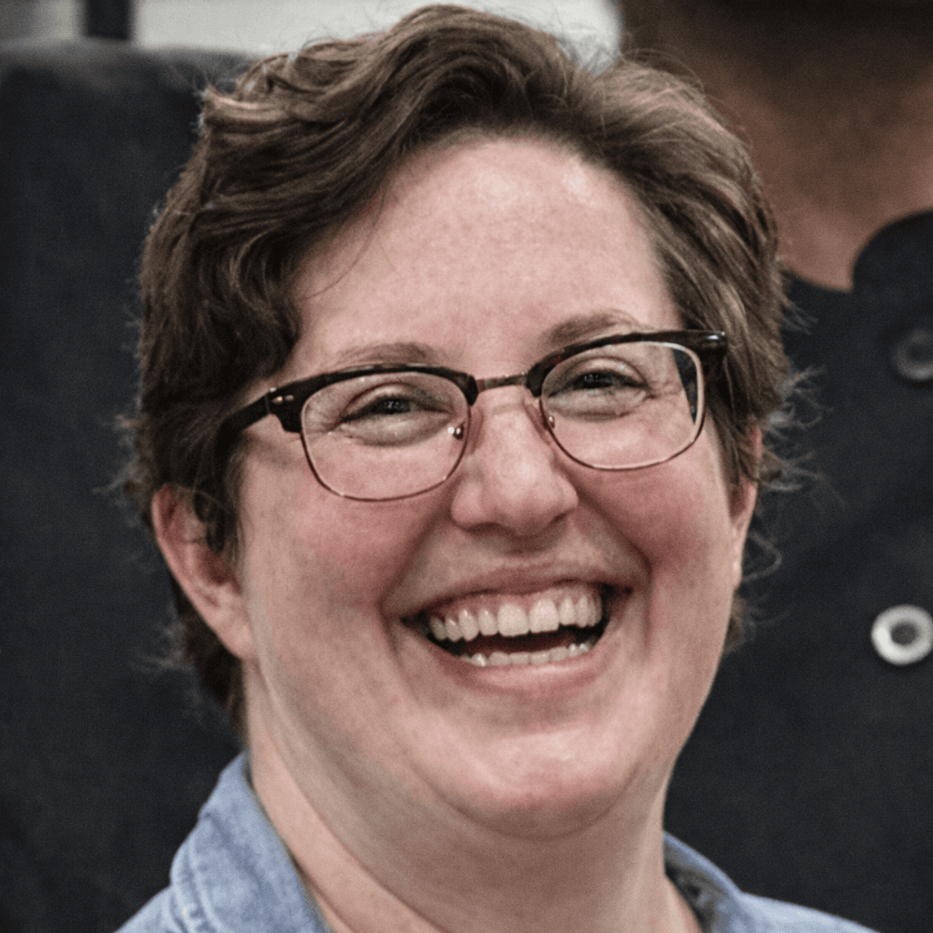 A person with short brown hair and glasses laughs joyfully in a close-up portrait.